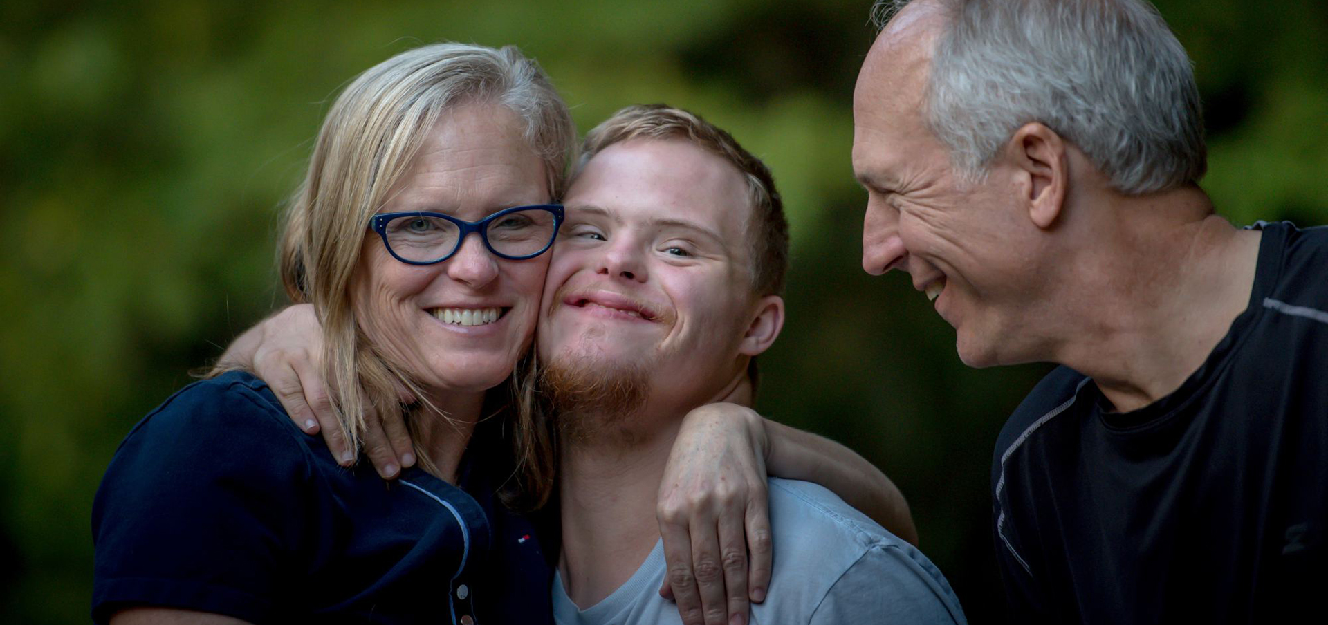 Eine dreiköpfige Familie lacht freundlich in die Kamera, sie nehmen ihren Sohn in den Arm, der an Trisomie 21 erkrankte. Der Mann links von ihm lächelt ihn freundlich an, während die Frau rechts von ihm ihn in den Arm nimmt.