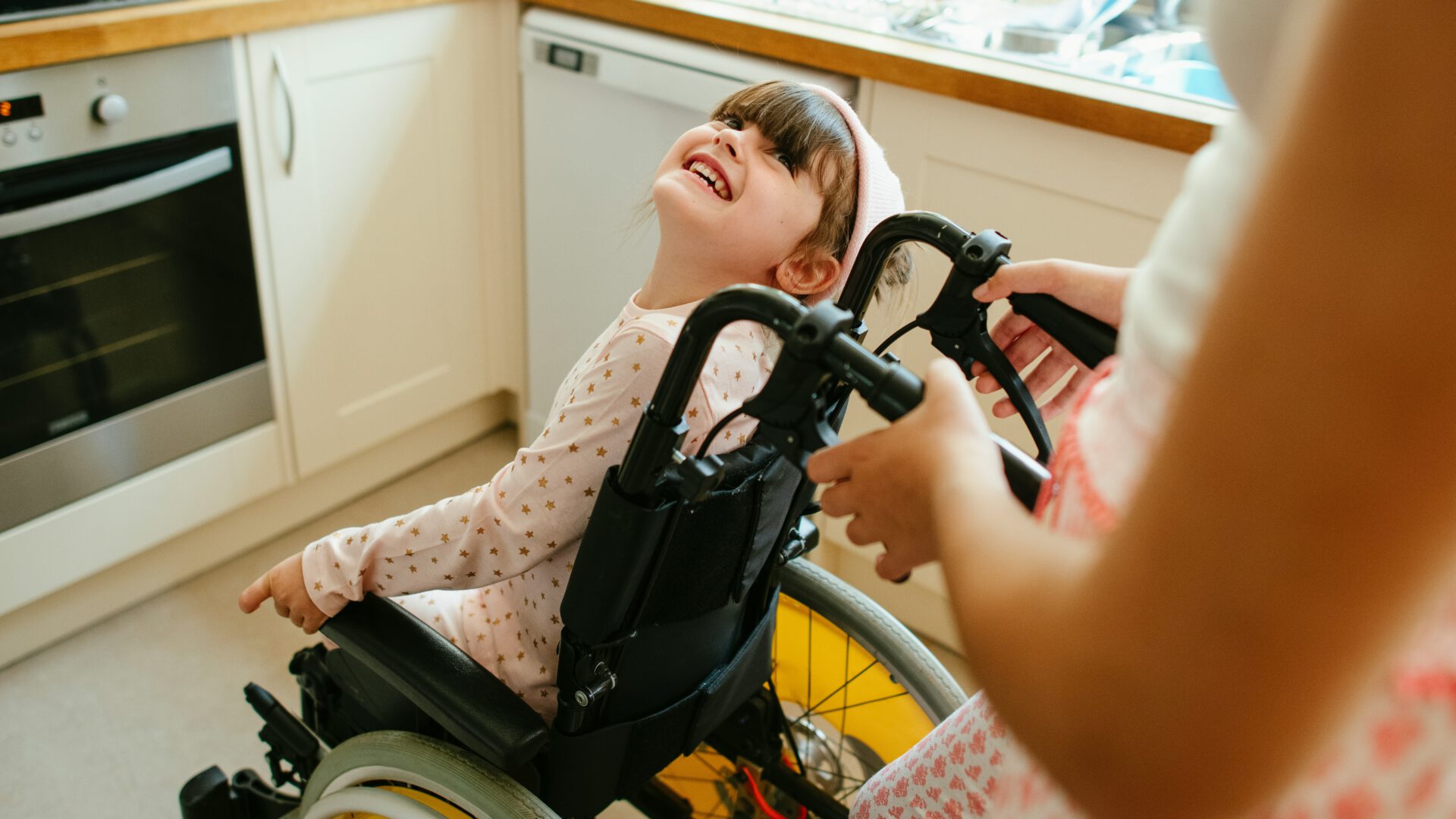 kleines Mädchen im Rollstuhl, steht in der Küche und schaut zu einer stehenden Person hoch, die nur verschwommen und abgeschnitten zu erkennen ist