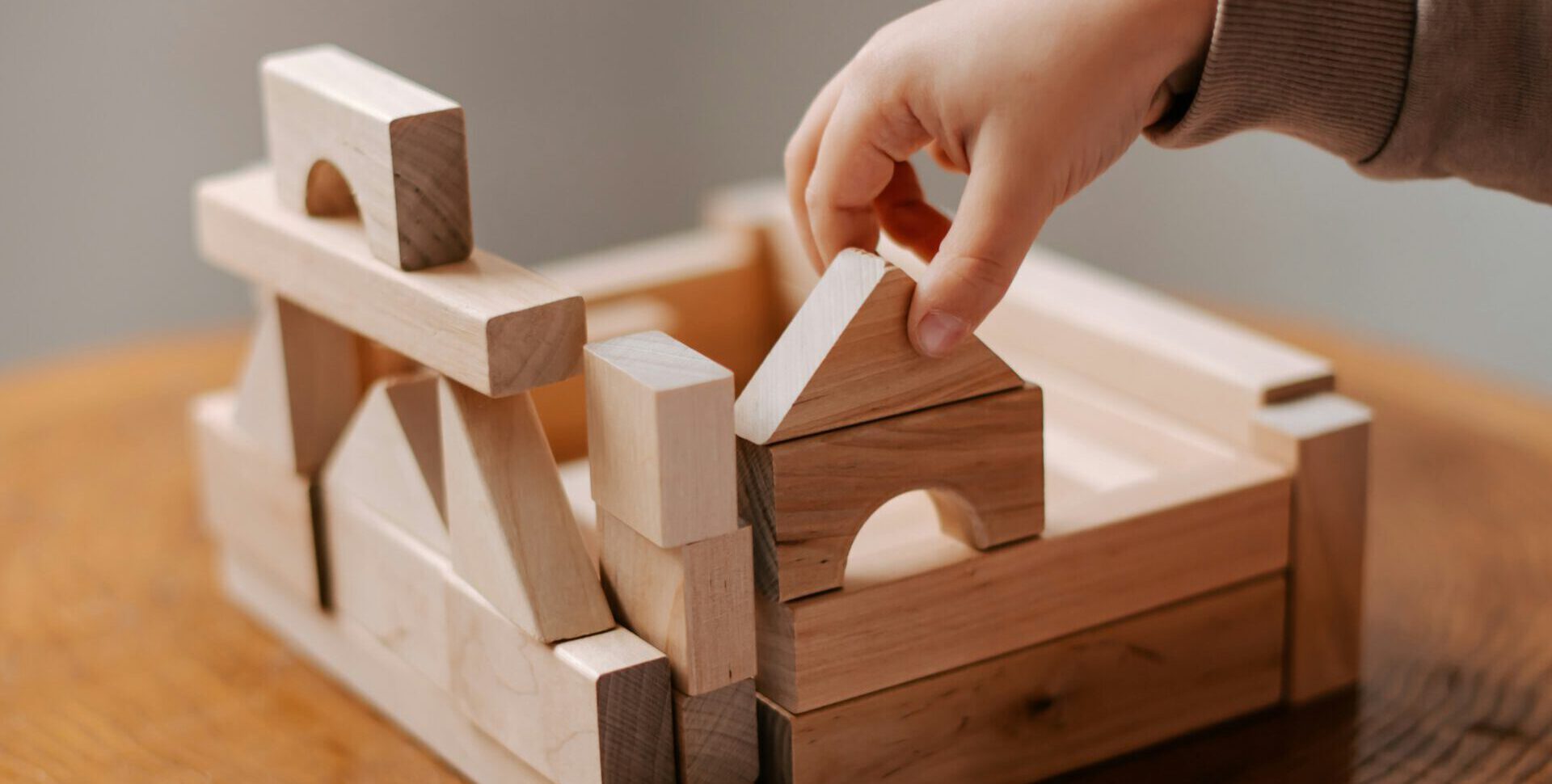 Kinderhand legt Holz-Baustein auf die anderen Bausteine, als Tisch ist ein Baumstumpf zu erkennen